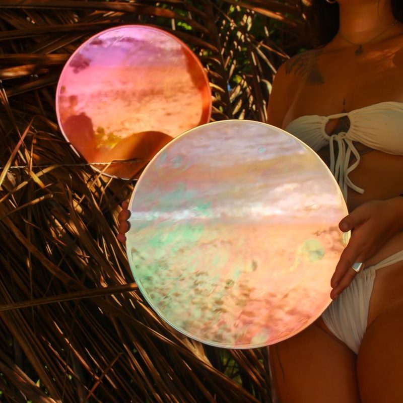 Dois espelhos redondos iridescentes, em rosa, laranja e verde, segurados entre folhas secas de palmeira.