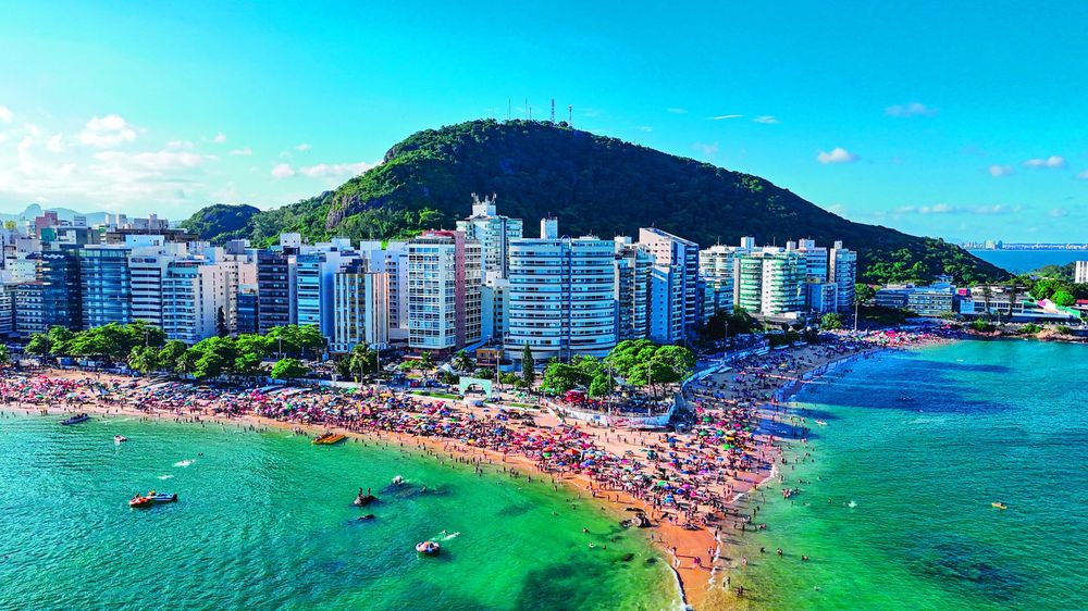 Vista aérea da Praia da Costa cheia de guarda-sóis, com os prédios da orla e o Morro do Moreno ao fundo.