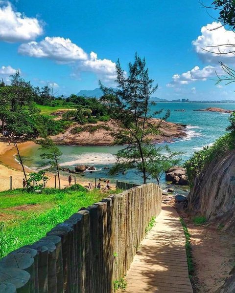 Caminho de madeira ao lado de uma cerca de troncos, descendo até uma pequena praia de águas verdes entre pedras e árvores.